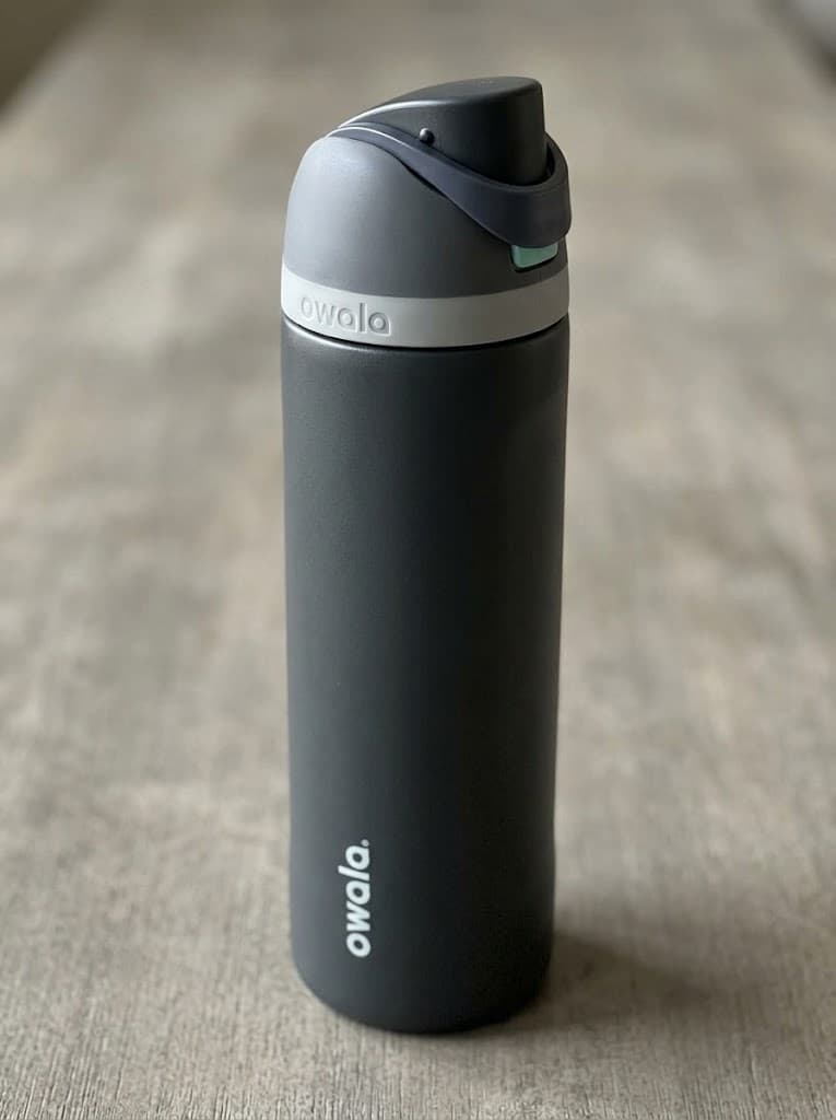 A dark Owala bottle with a gray lid band and carrying loop, standing on a neutral surface.
